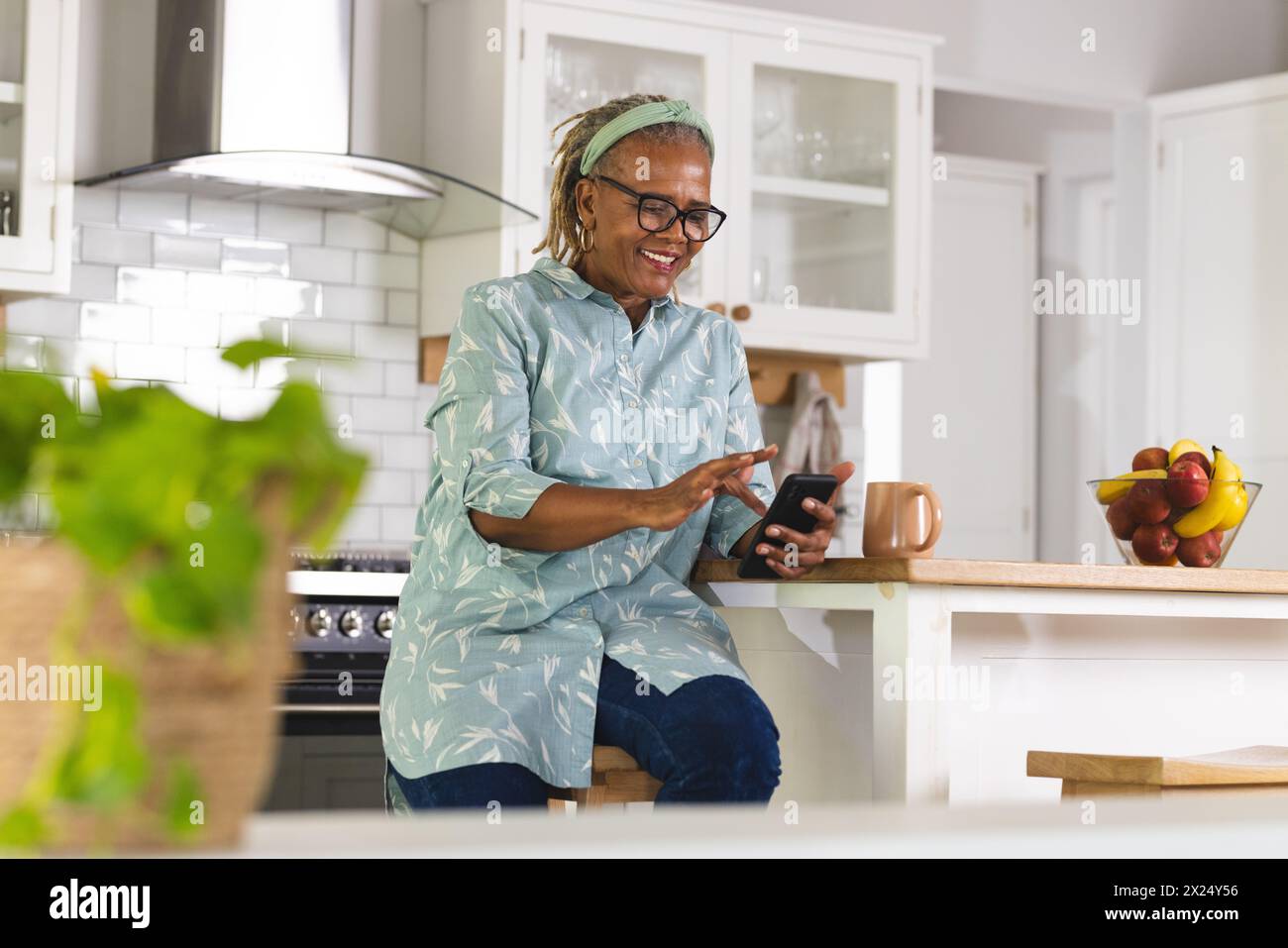 Une femme afro-américaine senior utilise un smartphone dans la cuisine à la maison, espace de copie. Elle a les cheveux gris courts, des lunettes, et porte un bleu clair Banque D'Images