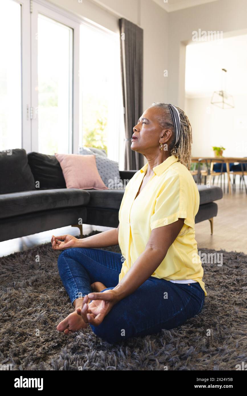 Femme afro-américaine senior, tresses grises, en chemise jaune, médite à la maison. Elle a fermé les yeux, assise les jambes croisées sur un tapis gris, inaltérée Banque D'Images