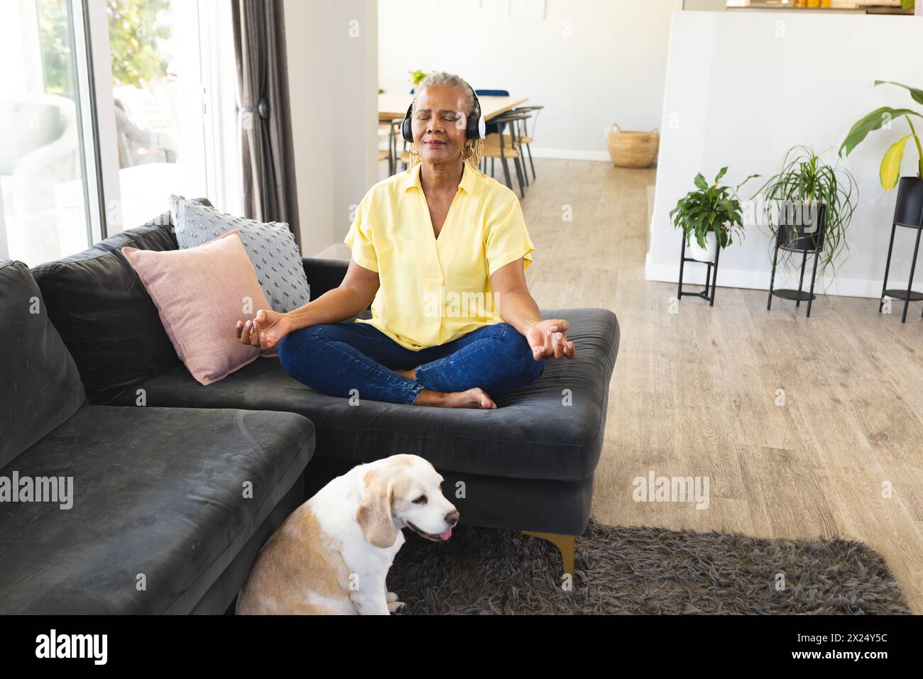 Une femme afro-américaine senior assise les jambes croisées, méditant sur un canapé à la maison. Elle a les cheveux gris courts, portant une chemise jaune et un Jean bleu, wi Banque D'Images