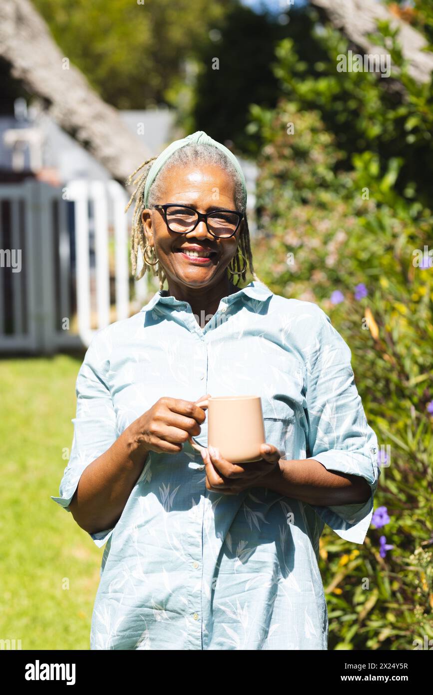 Femme aînée afro-américaine tenant une tasse, debout dans un jardin ensoleillé à la maison Banque D'Images