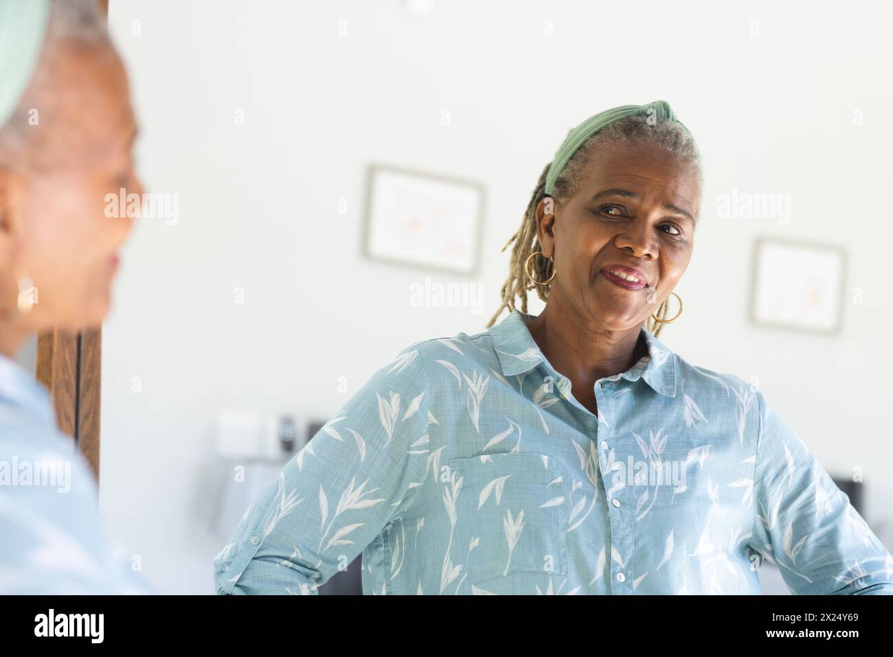 Une femme afro-américaine âgée avec les cheveux gris regardant dans le miroir à la maison. Vêtue d'une chemise bleu clair, elle affiche un sourire chaleureux, avec du fram en bois Banque D'Images