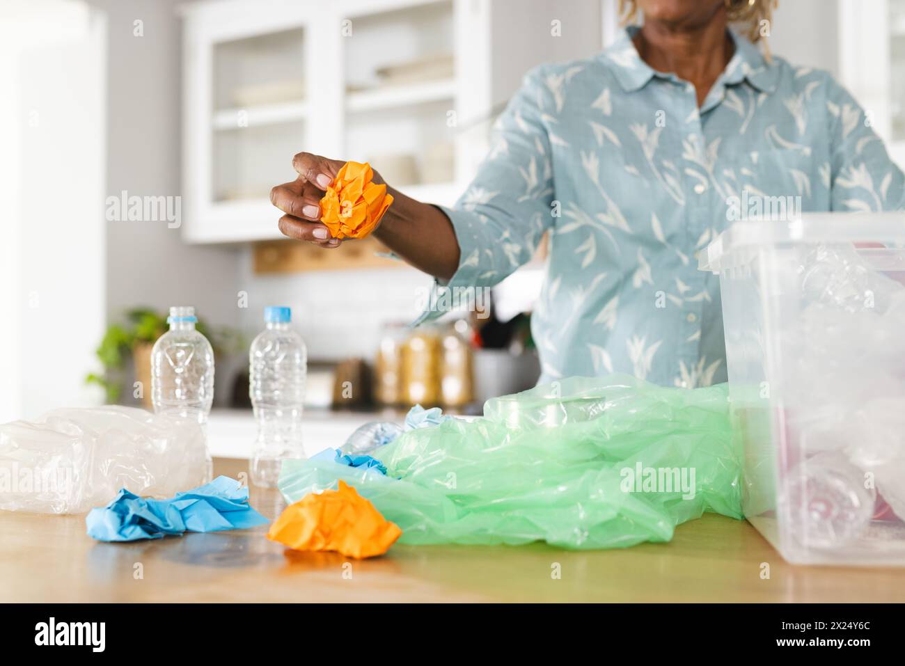 Une femme afro-américaine sénior triant des déchets plastiques pour les recycler à la maison. Elle a les cheveux courts gris et porte une chemise à motifs clairs, inaltérée. Banque D'Images
