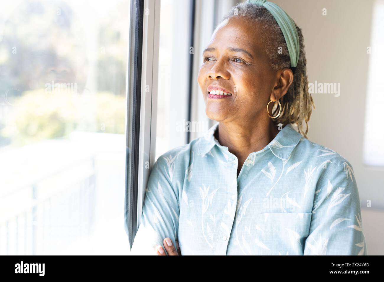 Une femme afro-américaine senior regarde dehors, debout près de la fenêtre à la maison, copie l'espace. Elle a les cheveux gris, les rides légères, et porte un Li Banque D'Images