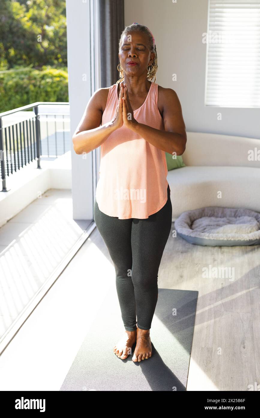 Une femme afro-américaine senior aux cheveux gris pratique le yoga à la maison. Elle porte un haut rose, des leggings noirs et se tient pieds nus sur un tapis. Banque D'Images
