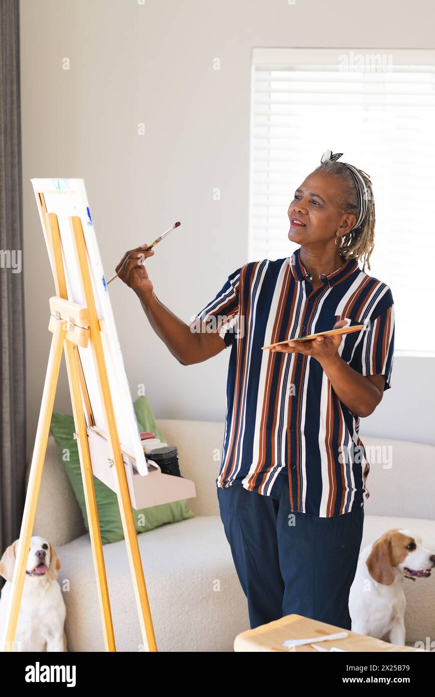 Femme aînée afro-américaine peignant sur toile à la maison, chiens regardant Banque D'Images