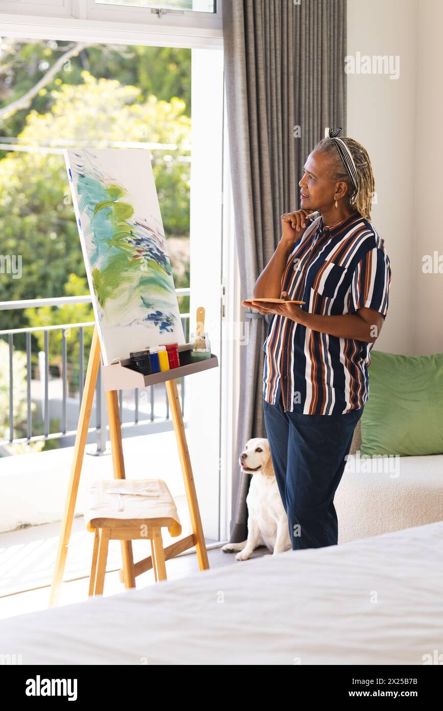 Femme afro-américaine senior avec des tresses grises en chemise rayée voit l'art à la maison Banque D'Images