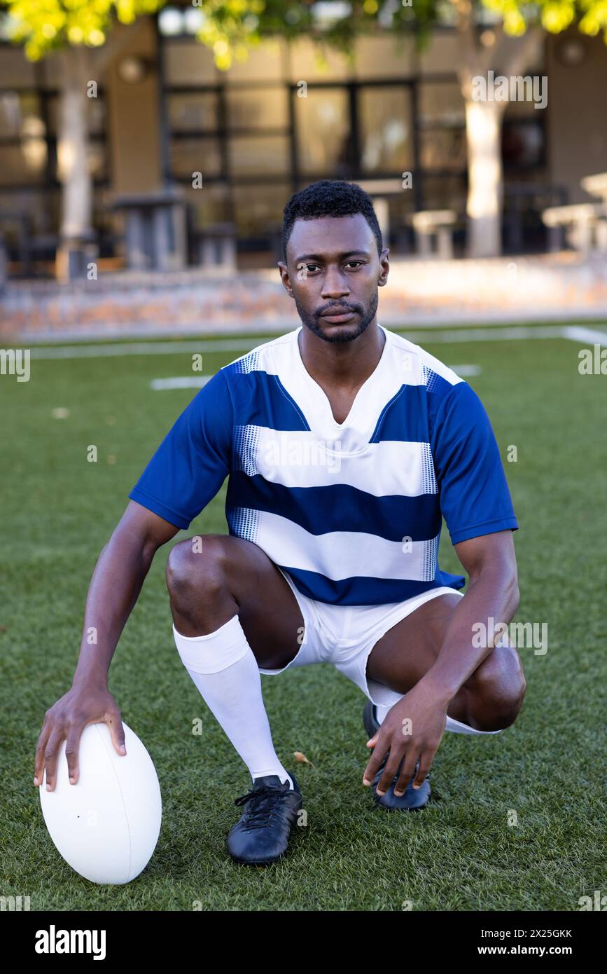 Jeune athlète afro-américain agenouillé sur le terrain à l'extérieur, tenant un ballon de rugby Banque D'Images
