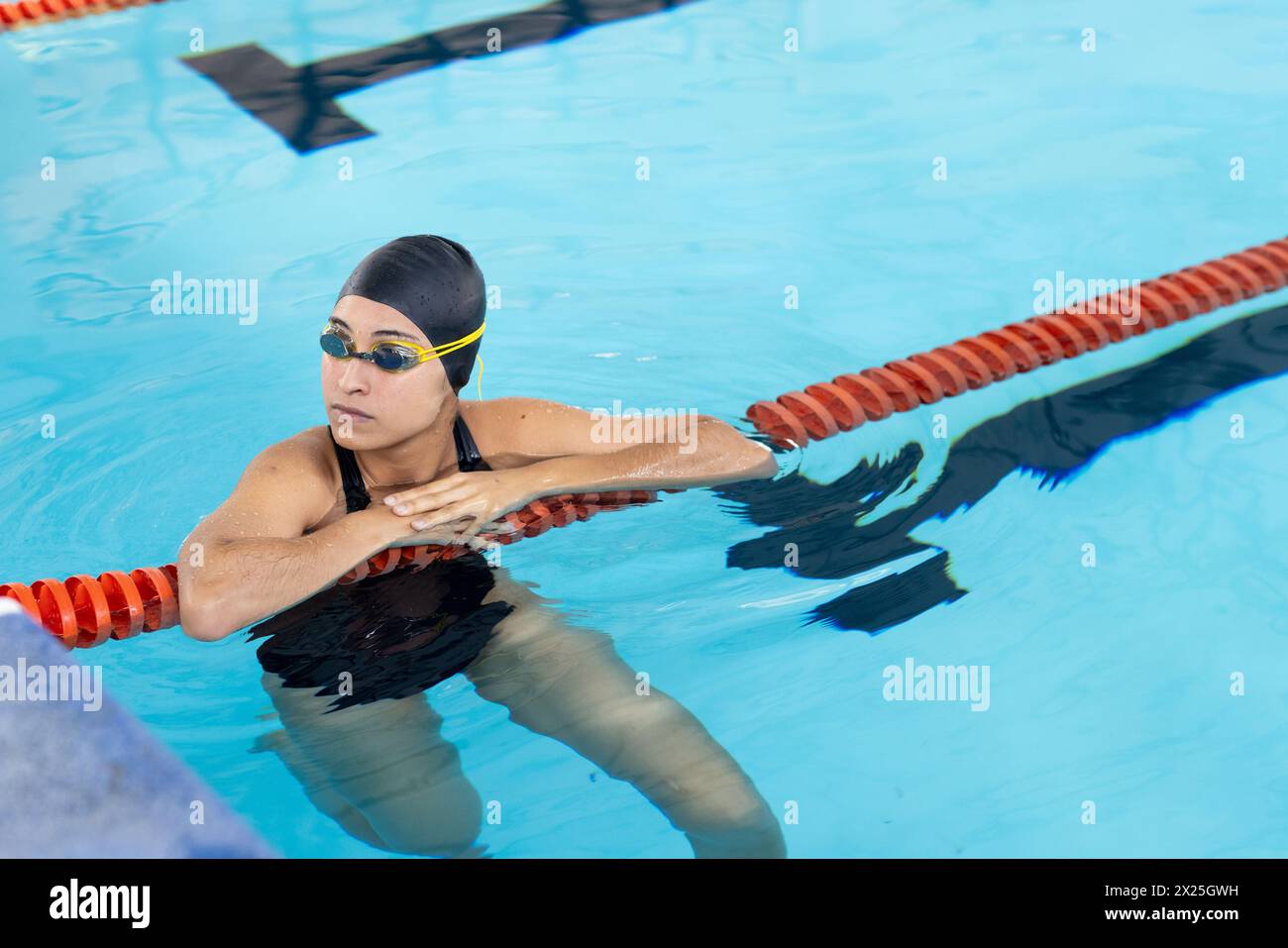 Jeune nageuse biraciale se reposant au bord de la piscine à l'intérieur, portant des lunettes de protection Banque D'Images