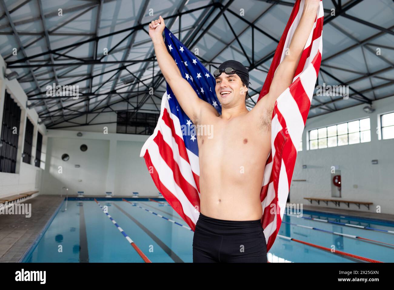 Jeune nageur caucasien tenant le drapeau américain, debout près de la piscine à l'intérieur, célébrant Banque D'Images