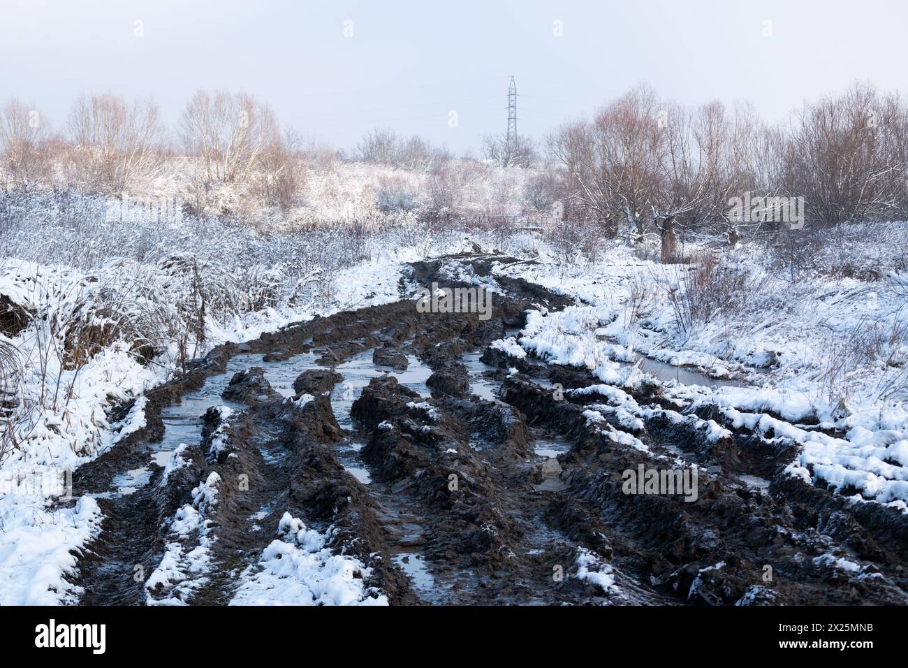 Traces de pneus sur route boueuse avec flaques d'eau glacées, paysage enneigé en hiver Banque D'Images