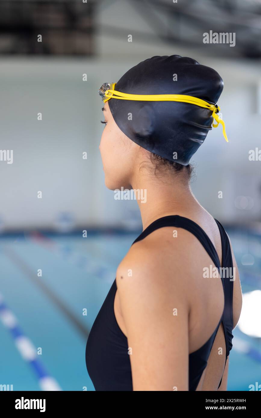 Jeune nageuse biraciale portant un maillot de bain noir et une casquette, debout au bord de la piscine à l'intérieur Banque D'Images