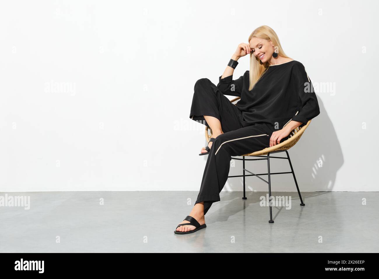 Femme blonde souriante en pyjama noir et sandales sur chaise Banque D'Images