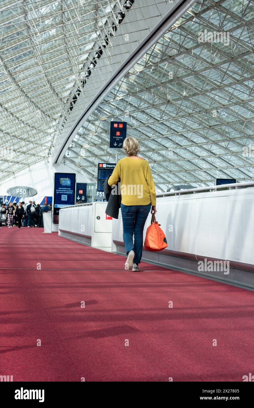 Vue arrière verticale d'une passager marchant avec sa valise à travers le terminal de l'aéroport de Paris pour monter à bord de son vol Banque D'Images