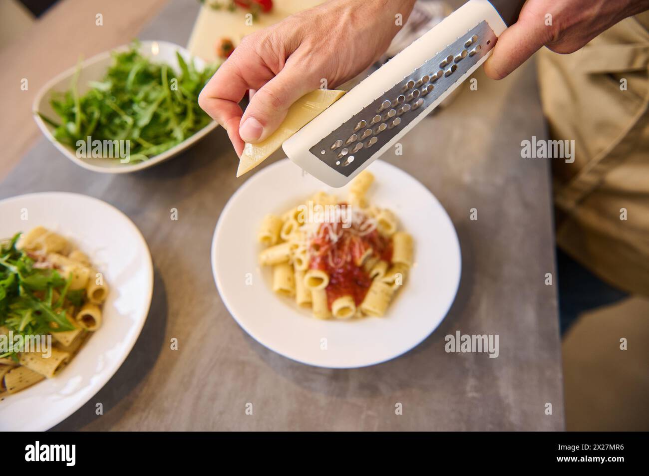 Directement au-dessus des mains du chef à l'aide d'une râpe, râper le fromage sur des pâtes fraîchement cuites avec de la sauce tomate, assaisonner et étaler le repas avant de le servir. Banque D'Images