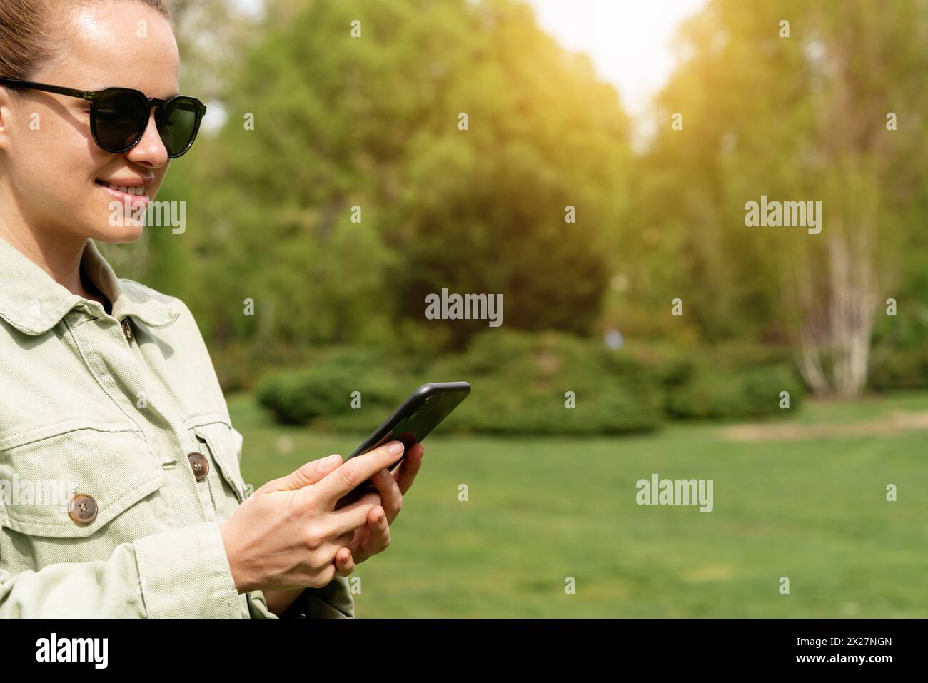 Femme dans des lunettes de soleil messagerie en ligne à l'aide d'un smartphone tout en marchant à l'extérieur dans un parc de la ville. Banque D'Images