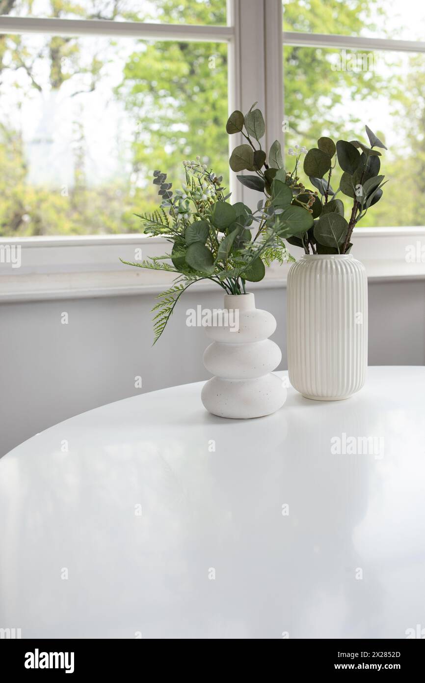 Fond de deux vases blancs avec verdure artificielle sur table blanche et fenêtre à l'arrière Banque D'Images