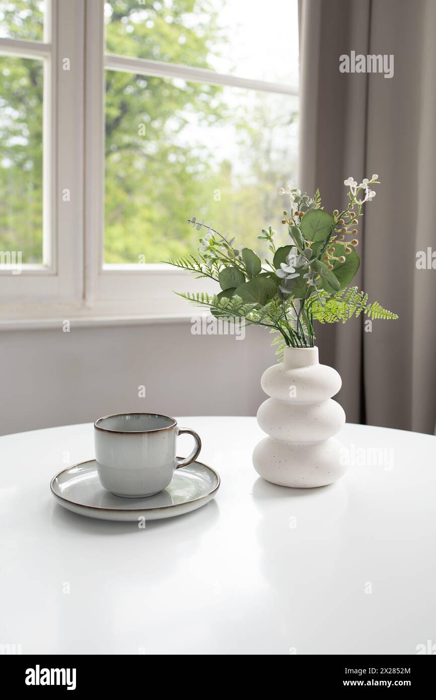 Vase blanc avec verdure artificielle et tasse vide sur table blanche et fenêtre à l'arrière Banque D'Images