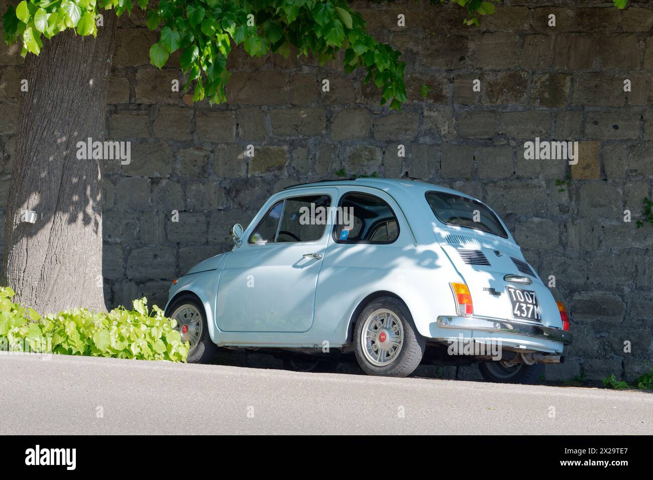 Bleu clair Classic Fiat 500 garé à côté d'un arbre et d'un haut mur de pierre à l'ombre à Montefiascone, Italie. 21 avril 2024 Banque D'Images