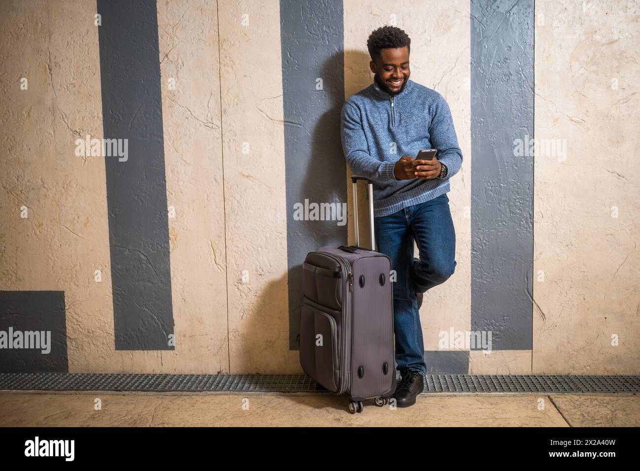 Homme heureux avec une valise en utilisant le téléphone tout en s'appuyant contre un mur dans le passage de la gare. Copiez l'espace sur l'image pour votre publicité ou votre texte. Banque D'Images
