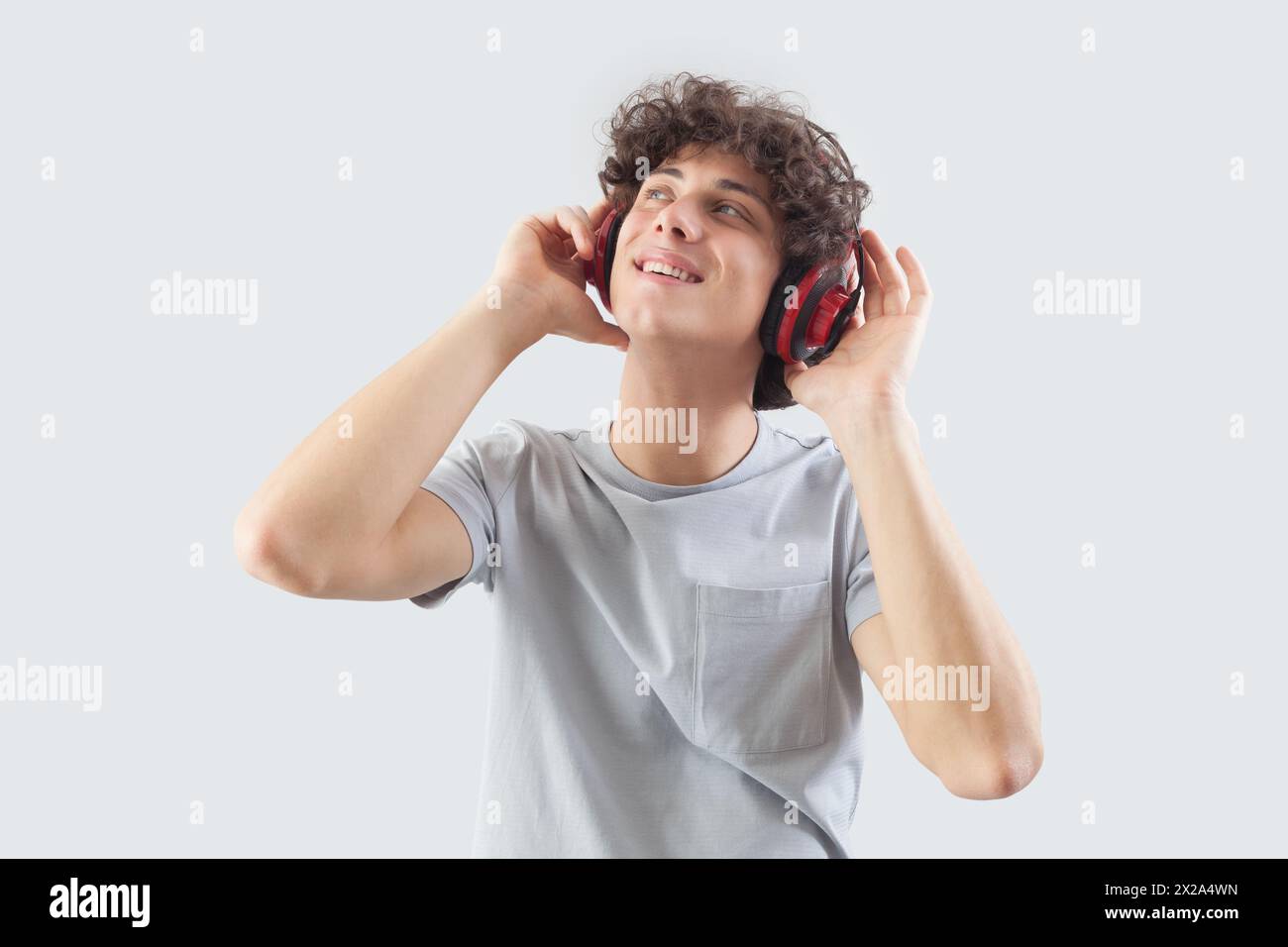 Souriant et beau, le jeune homme porte des écouteurs et est isolé sur un fond gris. Il regarde sur le côté avec ses yeux bleus tout en écoutant Banque D'Images