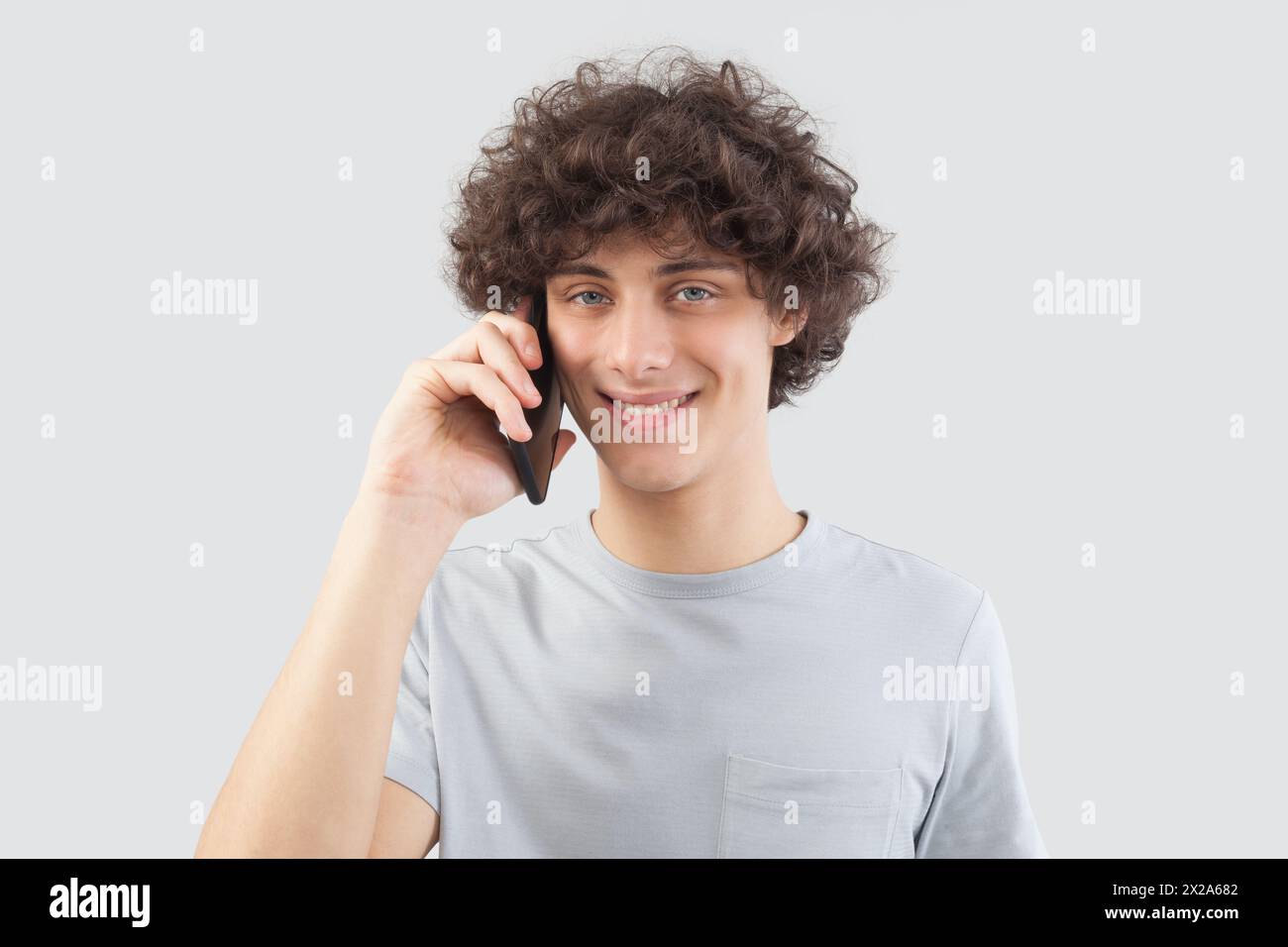 Un jeune homme beau sourit alors qu'il utilise son smartphone, parlant et écoutant avec son téléphone portable tout en regardant dans l'appareil photo avec ses yeux bleus. Il est moi Banque D'Images