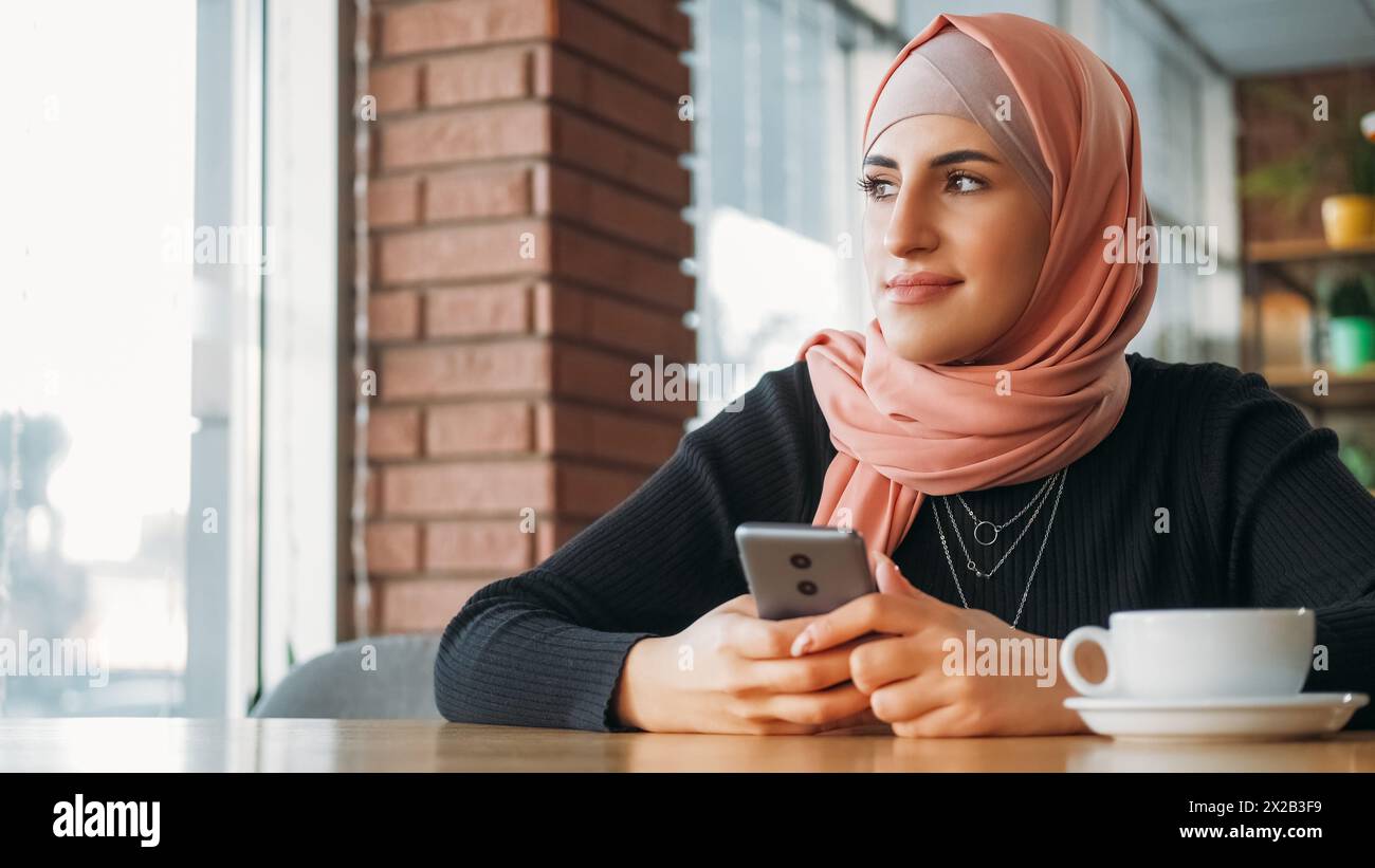 Mobile freelance. Loisirs gadgets. Femme souriante dans le hijab discutant sur le téléphone naviguant sur Internet assis à la fenêtre confortable du café. Banque D'Images