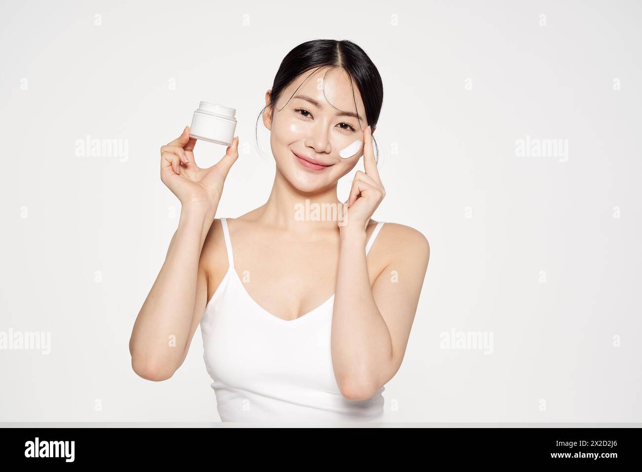 Une femme asiatique qui tient un récipient de crème et applique des cosmétiques sur son visage Banque D'Images