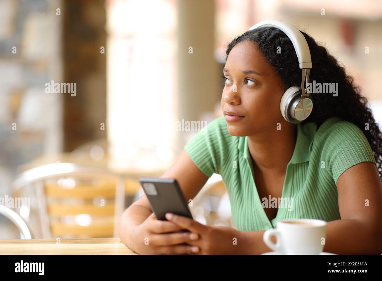 Femme noire pensant écouter l'audio avec casque et téléphone dans une terrasse de restaurant Banque D'Images