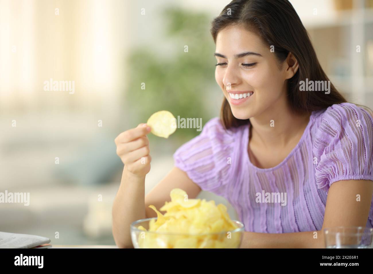 Femme heureuse mangeant des chips souriante assise à la maison Banque D'Images