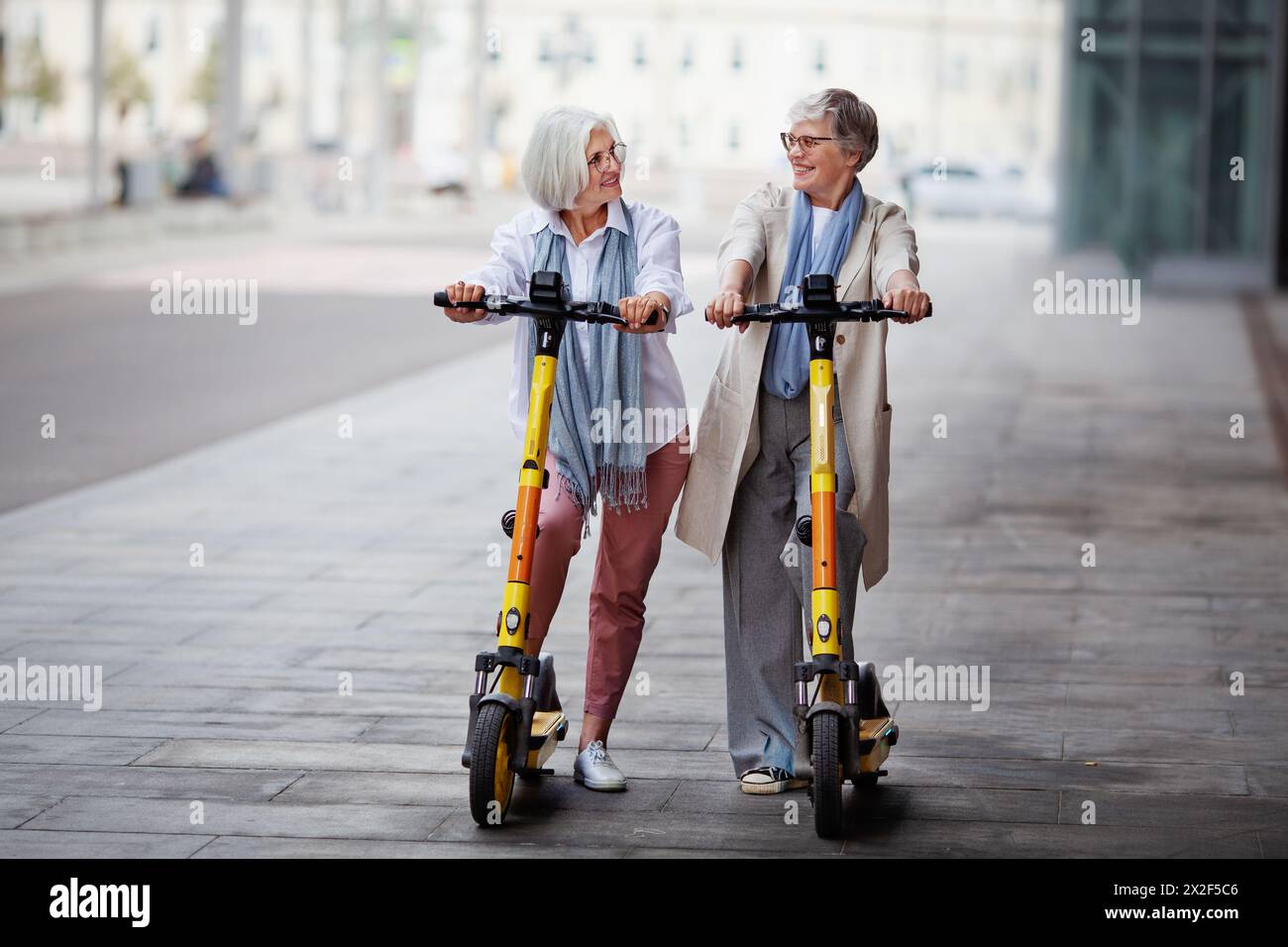 Matures joyeuses femmes aux cheveux gris amis montent des scooters électriques dans la ville, souriant heureux, profitant d'un moment amusant ensemble. Banque D'Images