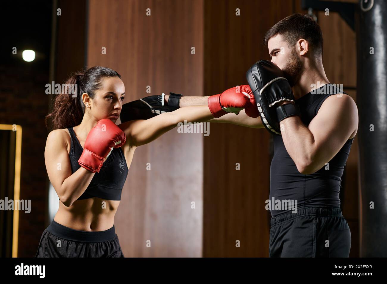 Un entraîneur masculin soutient une sportive brune en tenue active alors qu'elle s'engage dans l'entraînement de boxe dans un gymnase. Banque D'Images