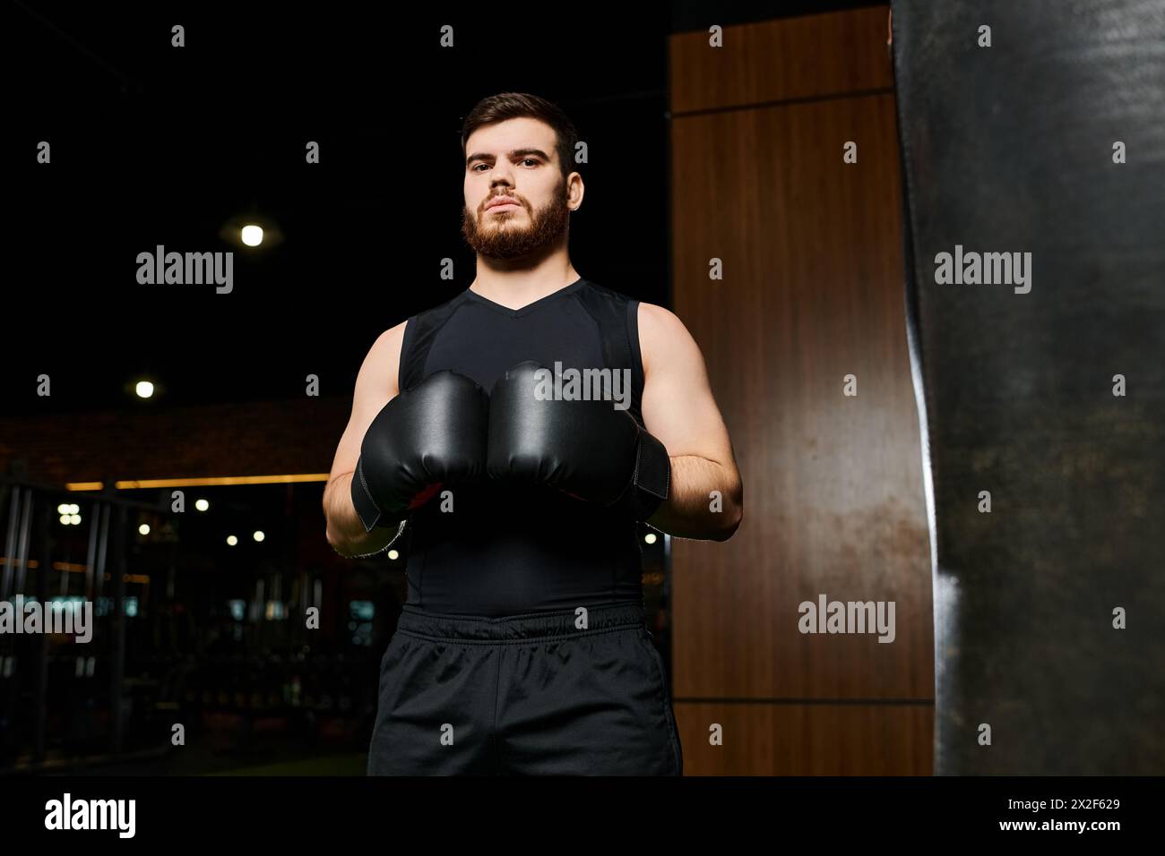 Un bel homme barbu portant des gants de boxe se tient confiant à côté d'un sac de frappe dans une salle de gym. Banque D'Images