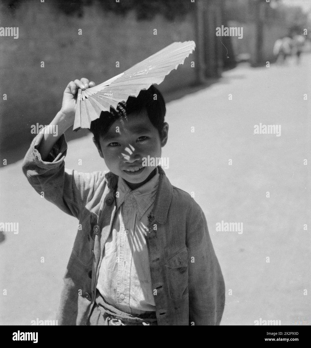 Un petit garçon de Chengtu, en Chine, pose avec un éventail tenu au-dessus de sa tête en 1944, illustrant les tenues traditionnelles, la vie quotidienne et la culture locale. Banque D'Images