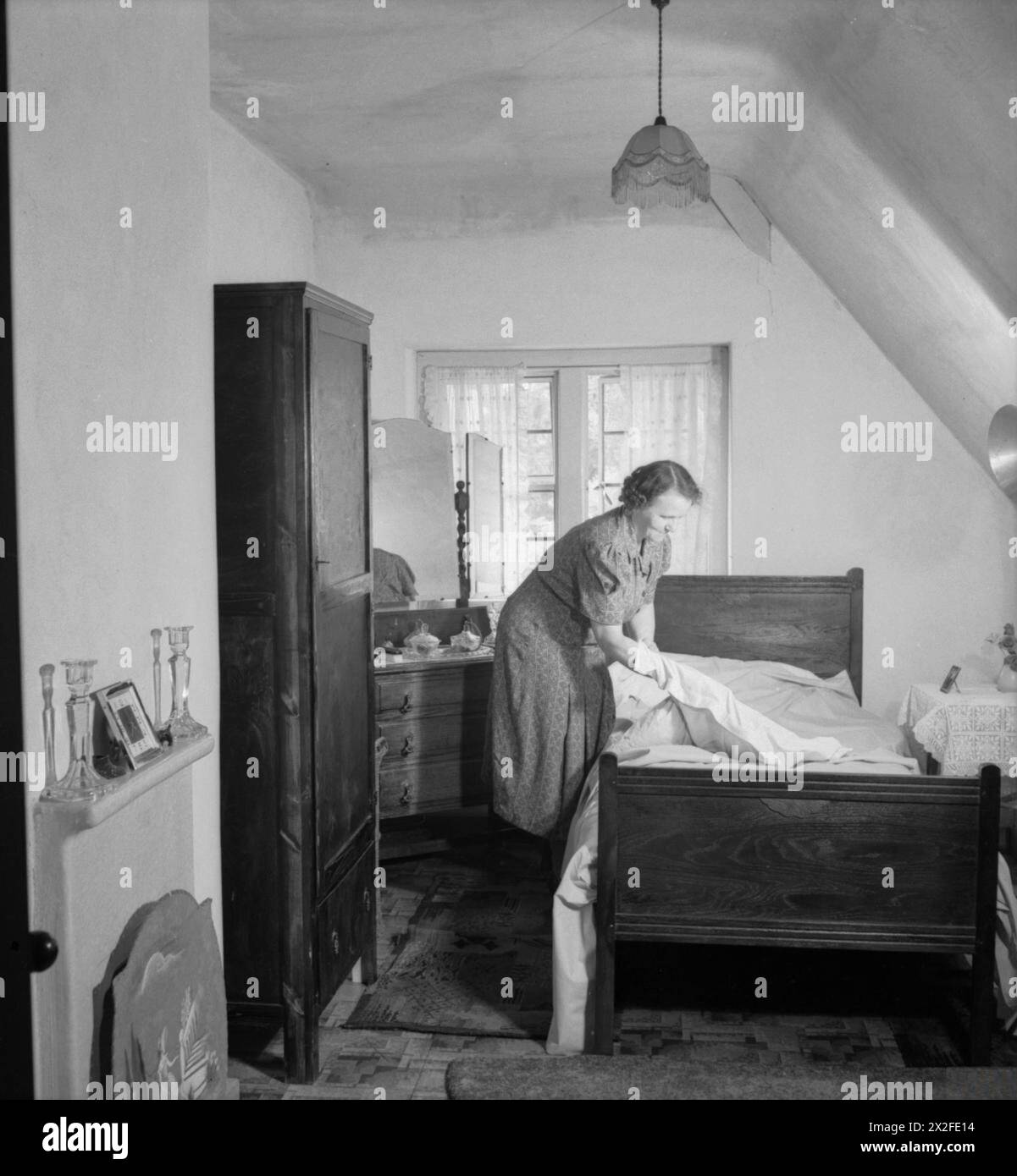 LES CHALETS DE FREEFOLK : LA VIE DANS LE VILLAGE DE FREEFOLK, HAMPSHIRE, ANGLETERRE, Royaume-Uni, 1943 - Mme Shadwell change le linge de lit dans l'une des chambres de sa maison dans le village de Freefolk dans le Hampshire. Chacun des salons et des chambres de ces maisons a une «cheminée carrelée moderne» (vue ici à gauche de la photo) et un éclairage électrique est installé dans toute la maison Banque D'Images