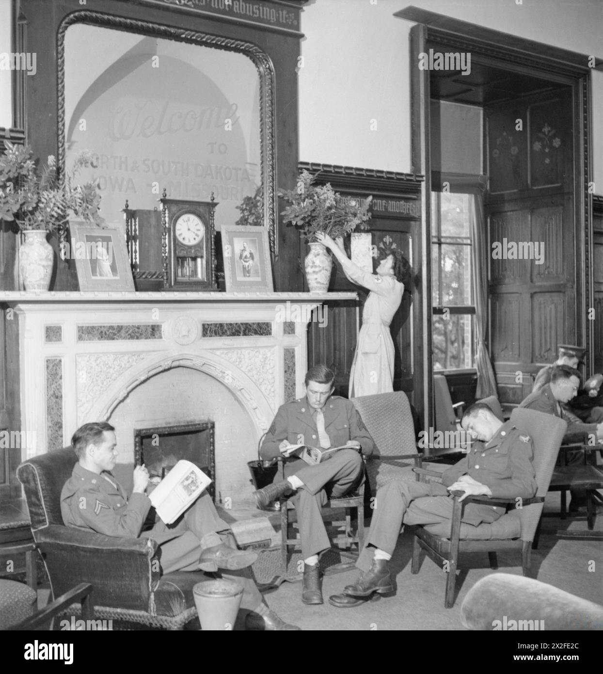 AMERICAN RED CROSS SERVICE CLUB, NORWICH : LA VIE AU CLUB AT THE BISHOP'S PALACE, NORFOLK, ANGLETERRE, ROYAUME-UNI, 1943 - Un membre du personnel de l'American Red Cross Service Club réarrange les fleurs sur la cheminée tandis que trois soldats américains se détendent dans des fauteuils devant le feu dans ce qui était autrefois la salle de dessin de Bishop, maintenant le salon du Club. Le caporal E William (à gauche) du 44 Glendale Road, Wellesley Hills, Massachusetts et le sergent Harold Martin (au centre) du 19 Willis Avenue, Floral Park, New York lisent les journaux, tandis qu'un troisième militaire dort. Le panneau au-dessus de la cheminée indique « Bienvenue au nord a » Banque D'Images