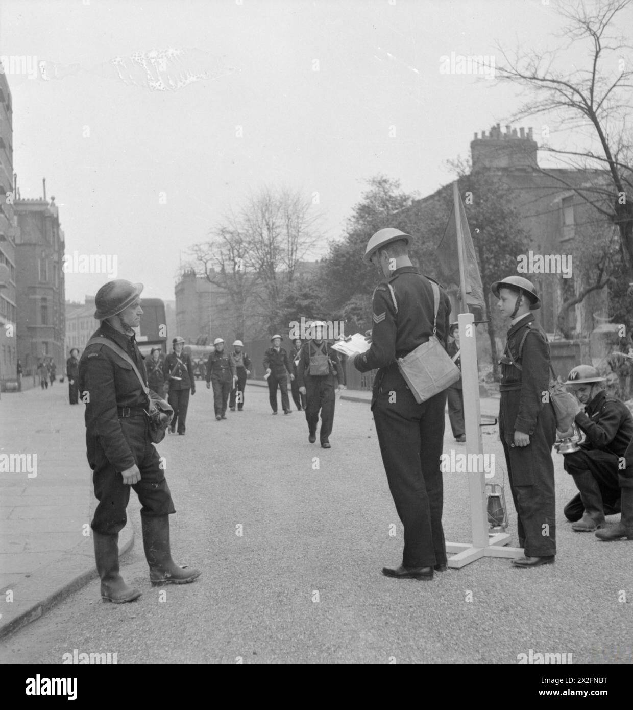 LA RECONSTRUCTION d'« UN INCIDENT » : FORMATION À LA DÉFENSE CIVILE À FULHAM, LONDRES, 1942 - les dirigeants des différents services arrivent et font rapport à l'officier responsable de l'incident. Sur la gauche, on peut voir le directeur de l'ARP qui a signalé l'incident à l'origine. L'officier en charge de l'incident porte un casque bleu, une lampe bleue et un drapeau bleu, pour lui-même et pour le point de contrôle de l'incident clairement visible par tous les agents de la protection civile impliqués. Un messager garçon peut également être vu. Cette photographie a été prise sur North End Road, à la jonction avec Conan Street (maintenant West Cromwell Road), regardant vers le sud en direction de Fulham. Juste visible sur le l Banque D'Images