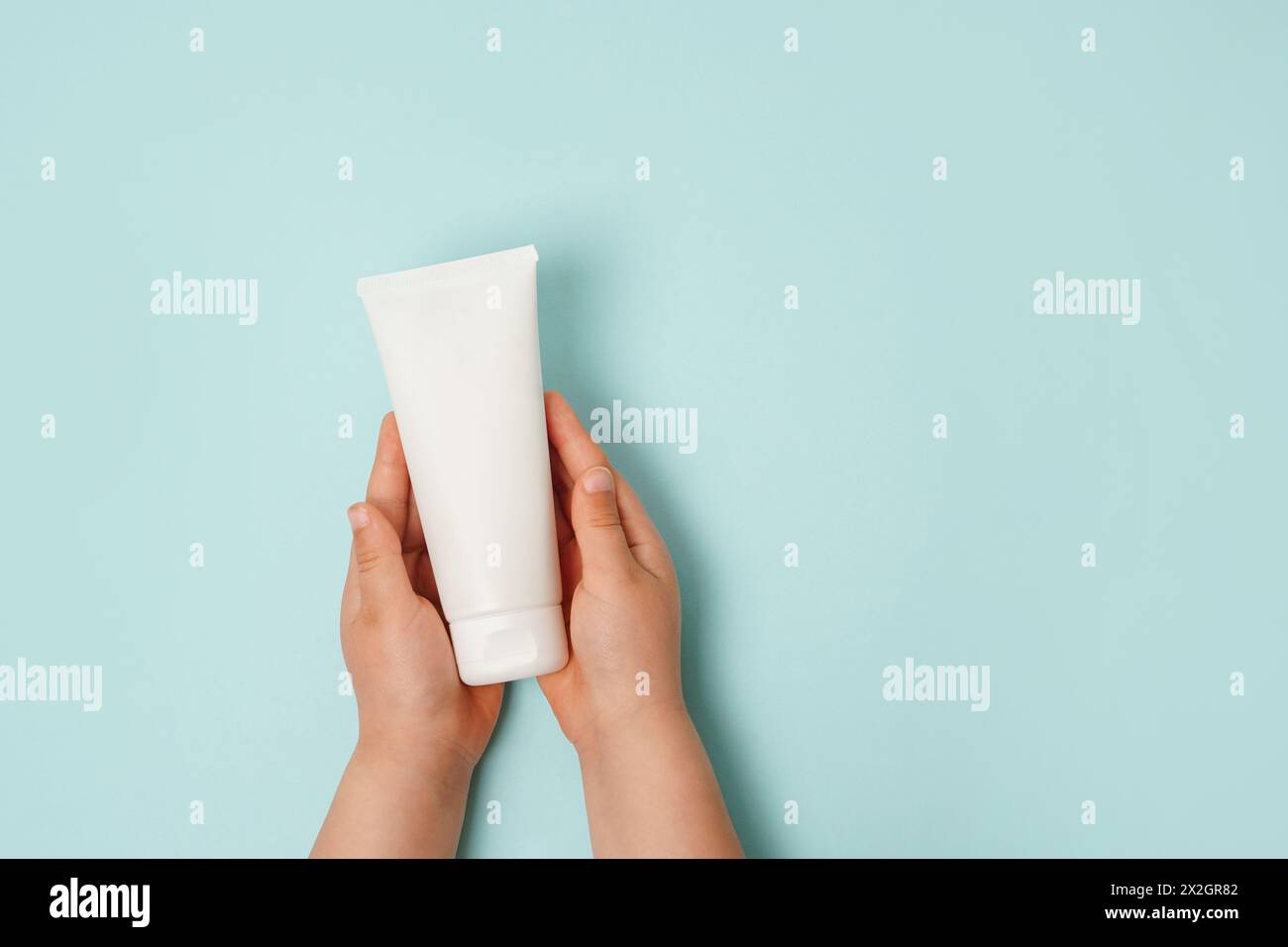 Mains d'enfant tenant le tube de crème blanche sur fond bleu clair. Attention à la peau propre et douce du corps. Produit de beauté quotidien pour enfants Banque D'Images