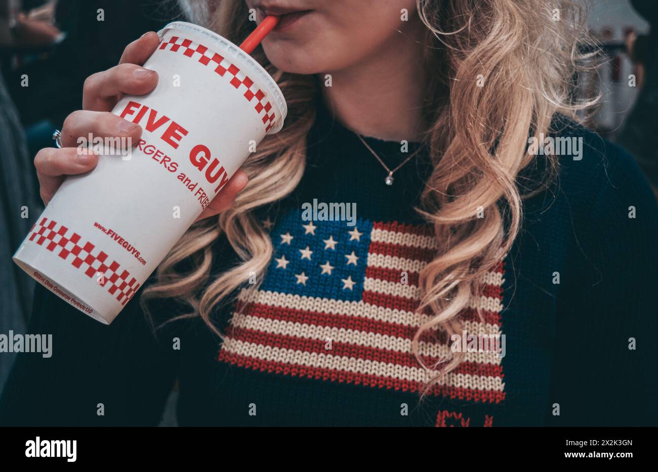 Gros plan d'une jeune femme sirotant une boisson dans une tasse de marque, portant un pull avec un motif de drapeau américain. Mettre l'accent sur les repas en plein air décontractés et le matin Banque D'Images