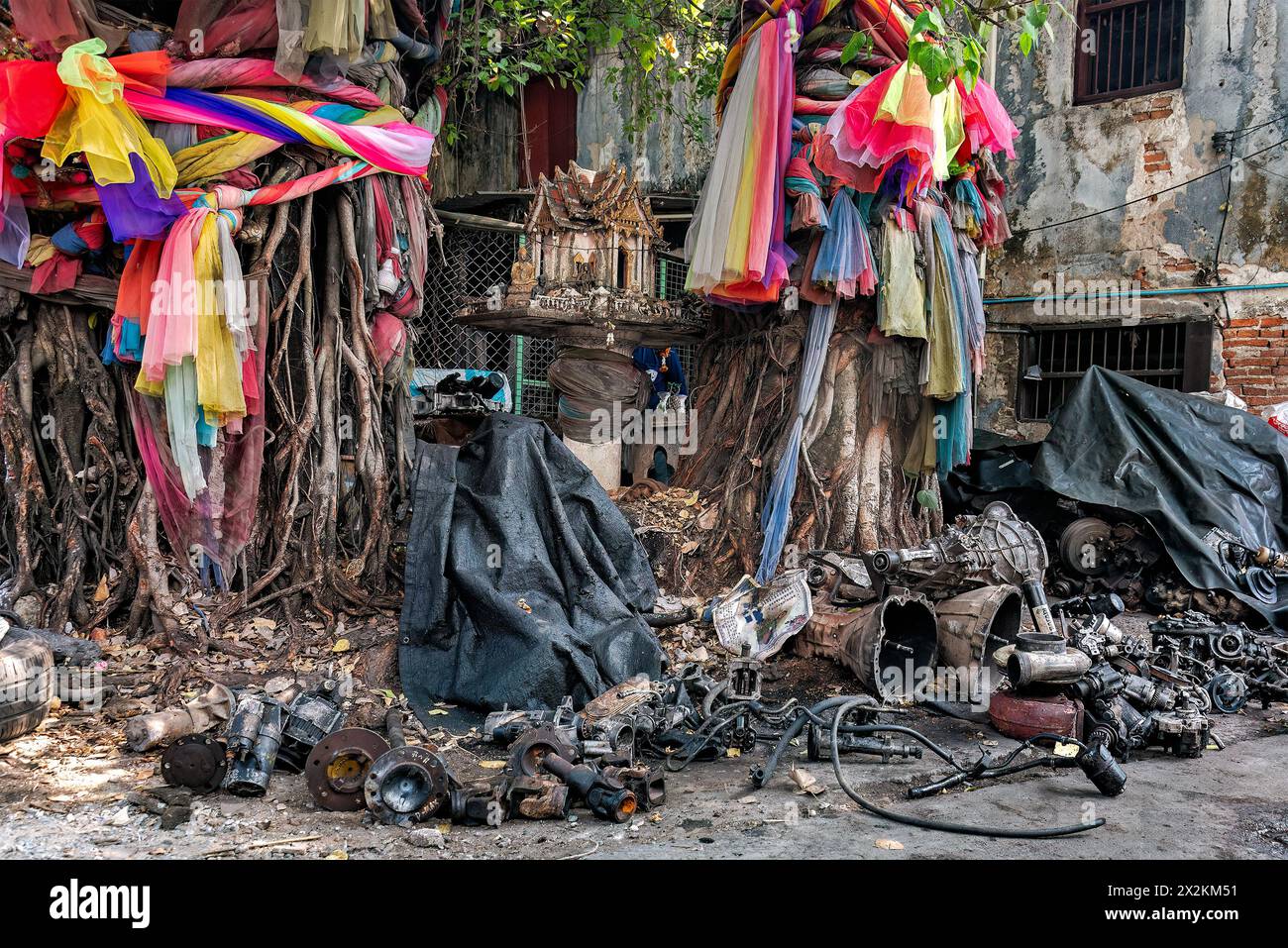 Les pièces de moteur de véhicule d'occasion sont laissées dispersées autour de 2 arbres bodhi attachés avec un assortiment de tissu coloré avec un sanctuaire Phra Phum au milieu. Banque D'Images