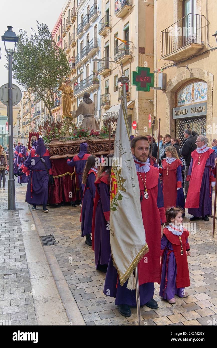 Tarragone, Espagne - 23 avril 2024 : procession traditionnelle avec des pénitents aux tons violets dans une rue pavée entourée de vieux bâtiments et spectateur Banque D'Images