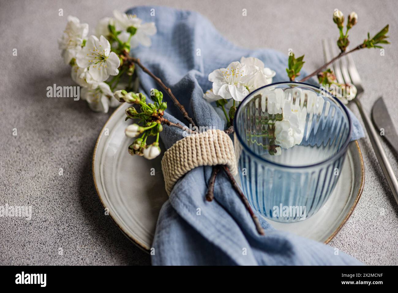 Une branche de fleur de cerisier fraîche complète une table sereine avec une assiette rustique, du verre rayé et un anneau de serviette sur un fond texturé Banque D'Images