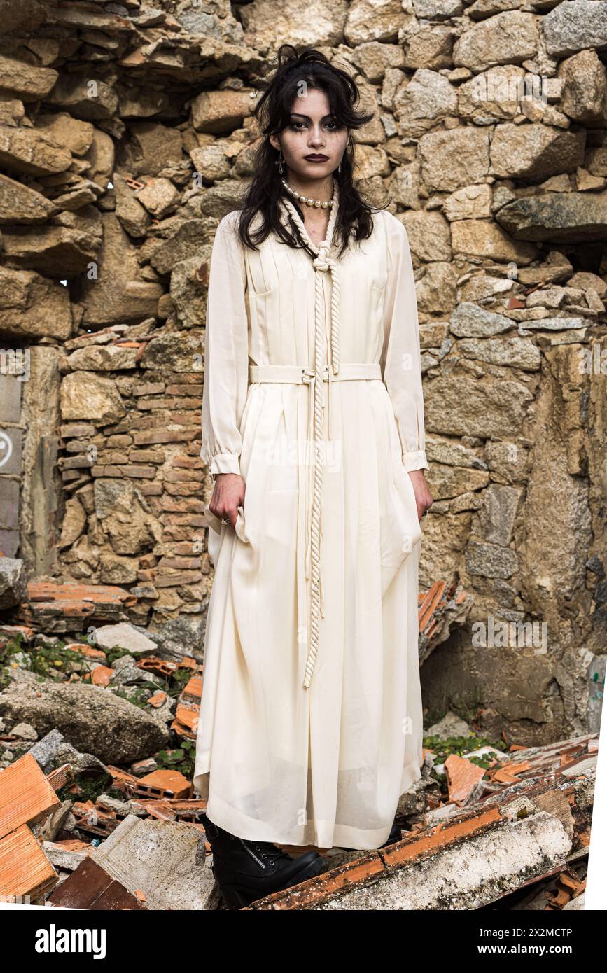 Une femme aux cheveux foncés et au maquillage se dresse dans une robe vintage crème devant un mur de pierre en ruine, dégageant une atmosphère gothique Banque D'Images