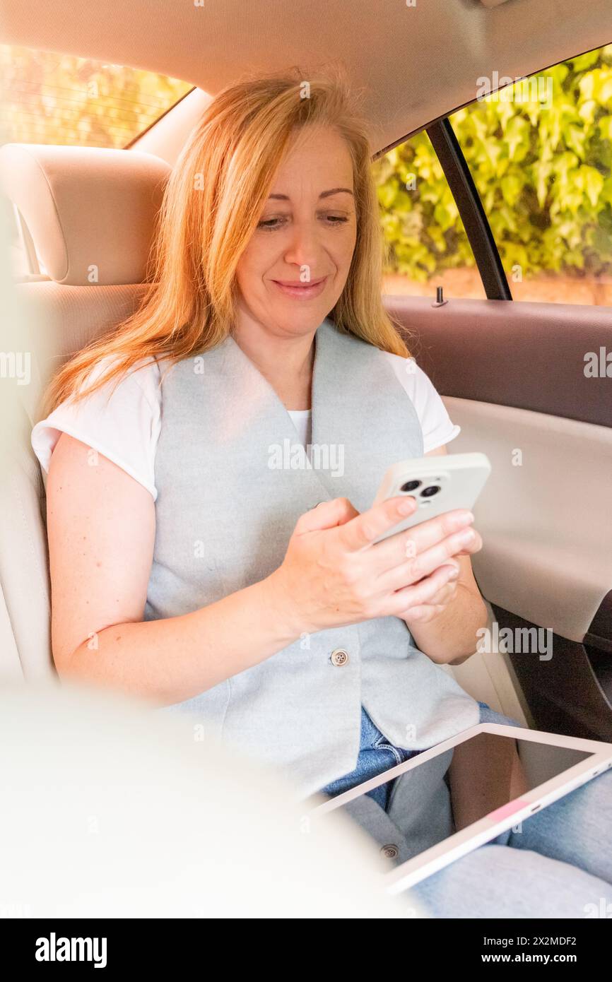 Femme souriante assise dans une voiture, utilisant son smartphone avec une tablette à côté d'elle Banque D'Images