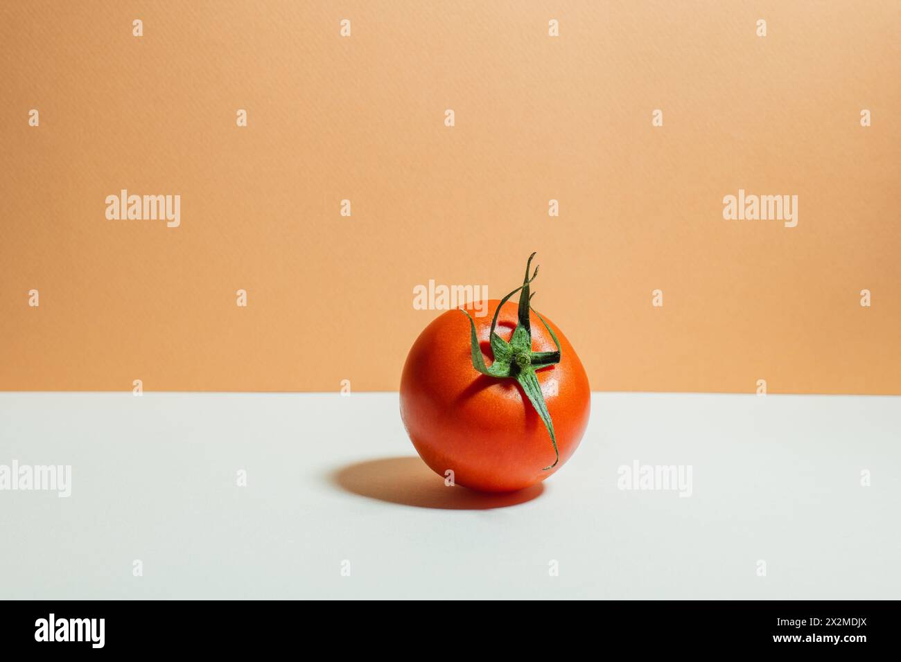 Une tomate rouge mûre avec une tige verte vibrante placée sur un fond bicolore blanc et orange, mettant en valeur la simplicité et la fraîcheur. Banque D'Images