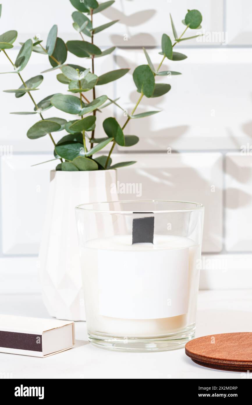 Une bougie parfumée contemporaine dans un récipient en verre transparent, accompagnée d'une plante d'eucalyptus dans un vase blanc, sur un fond carrelé blanc Banque D'Images