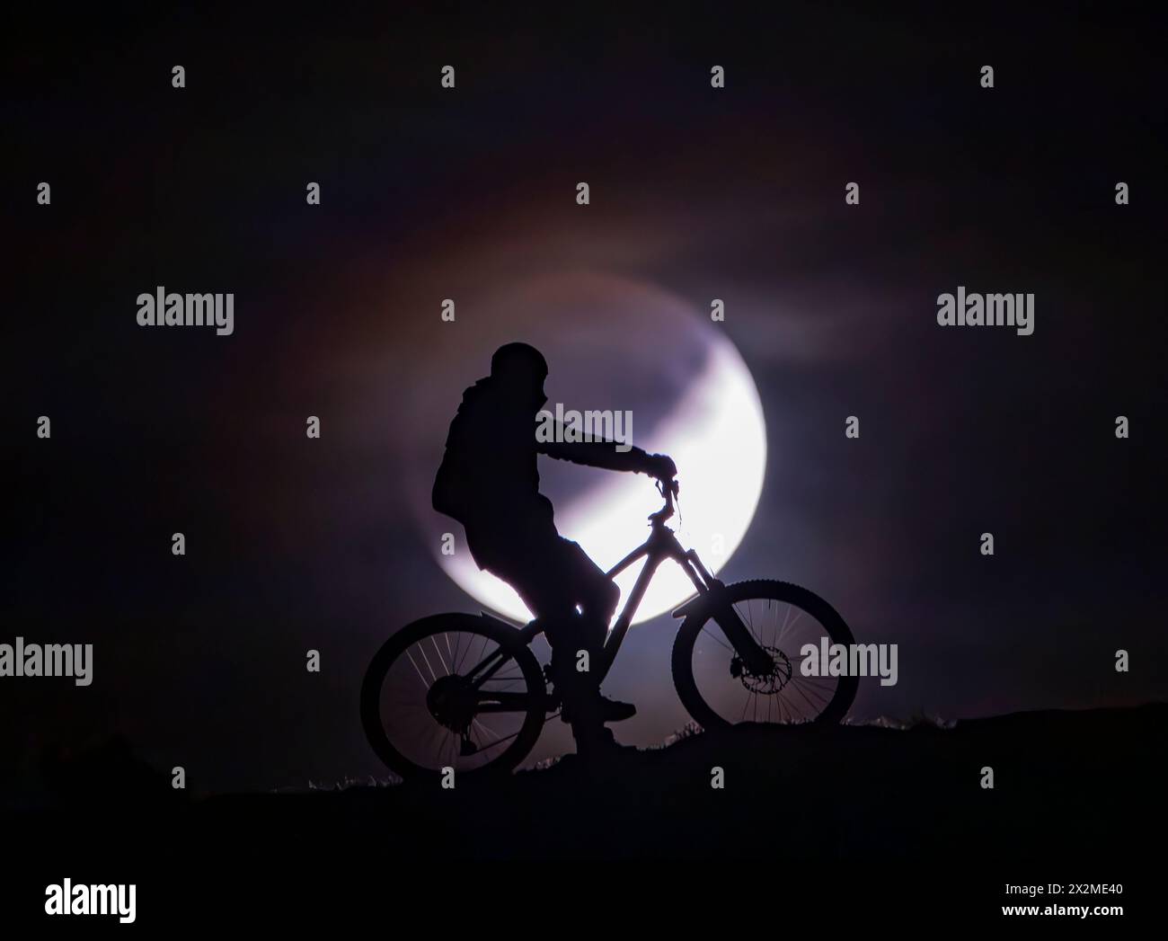 Silhouette d'un cycliste avec un VTT contre un ciel plein clair de lune, véhiculant l'aventure et l'esprit des balades nocturnes d'été Banque D'Images