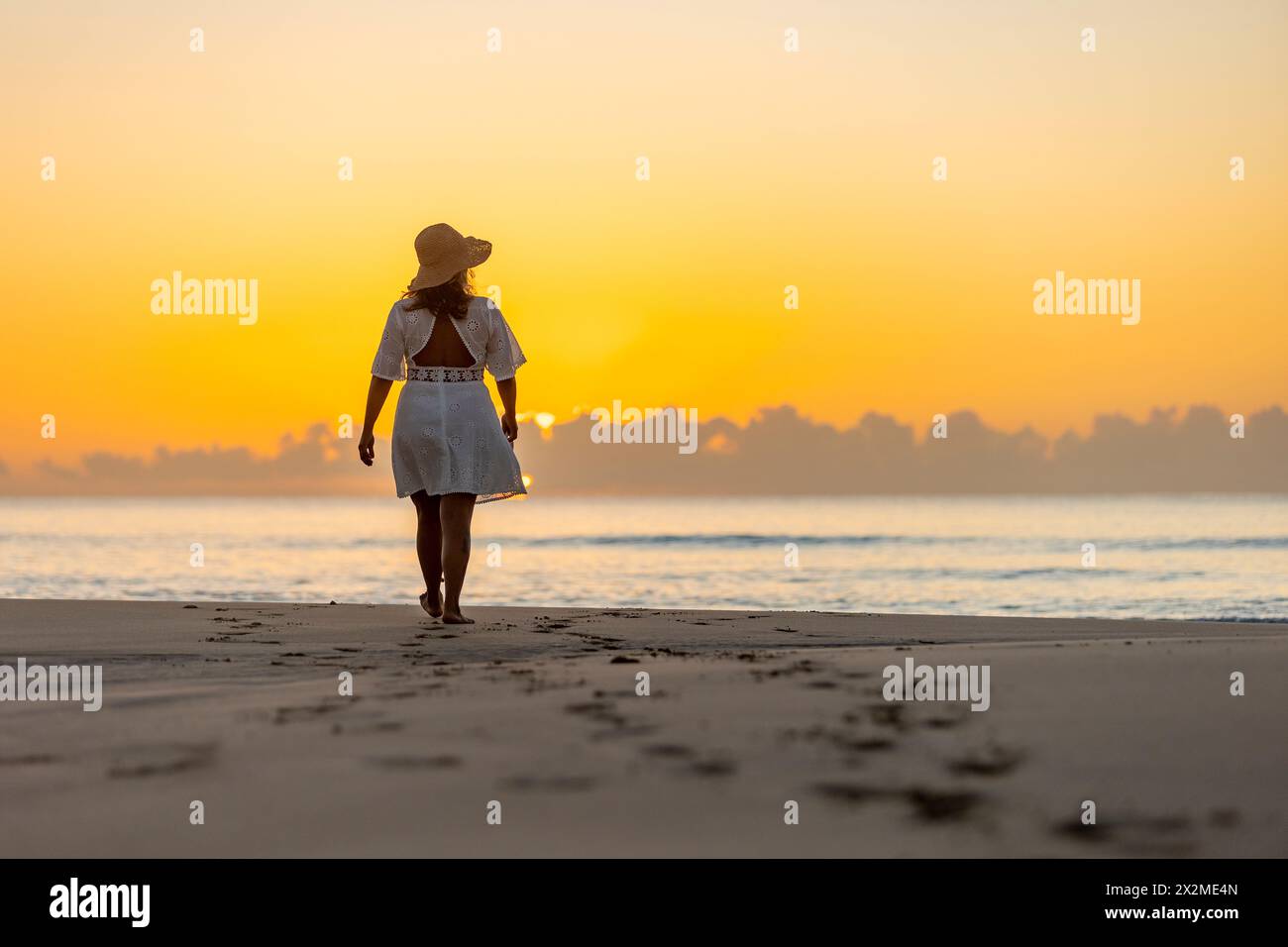 Vue arrière d'une femme méconnaissable profitant d'une promenade paisible le long du rivage au lever du soleil, avec la lueur chaude du soleil jetant une toile de fond tranquille. Banque D'Images