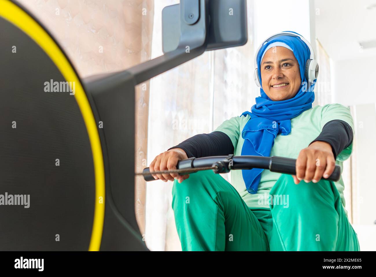 Une femme arabe dans un hijab et un casque sourit tout en s'entraînant sur des équipements de gymnastique, illustrant la diversité dans la forme physique Banque D'Images