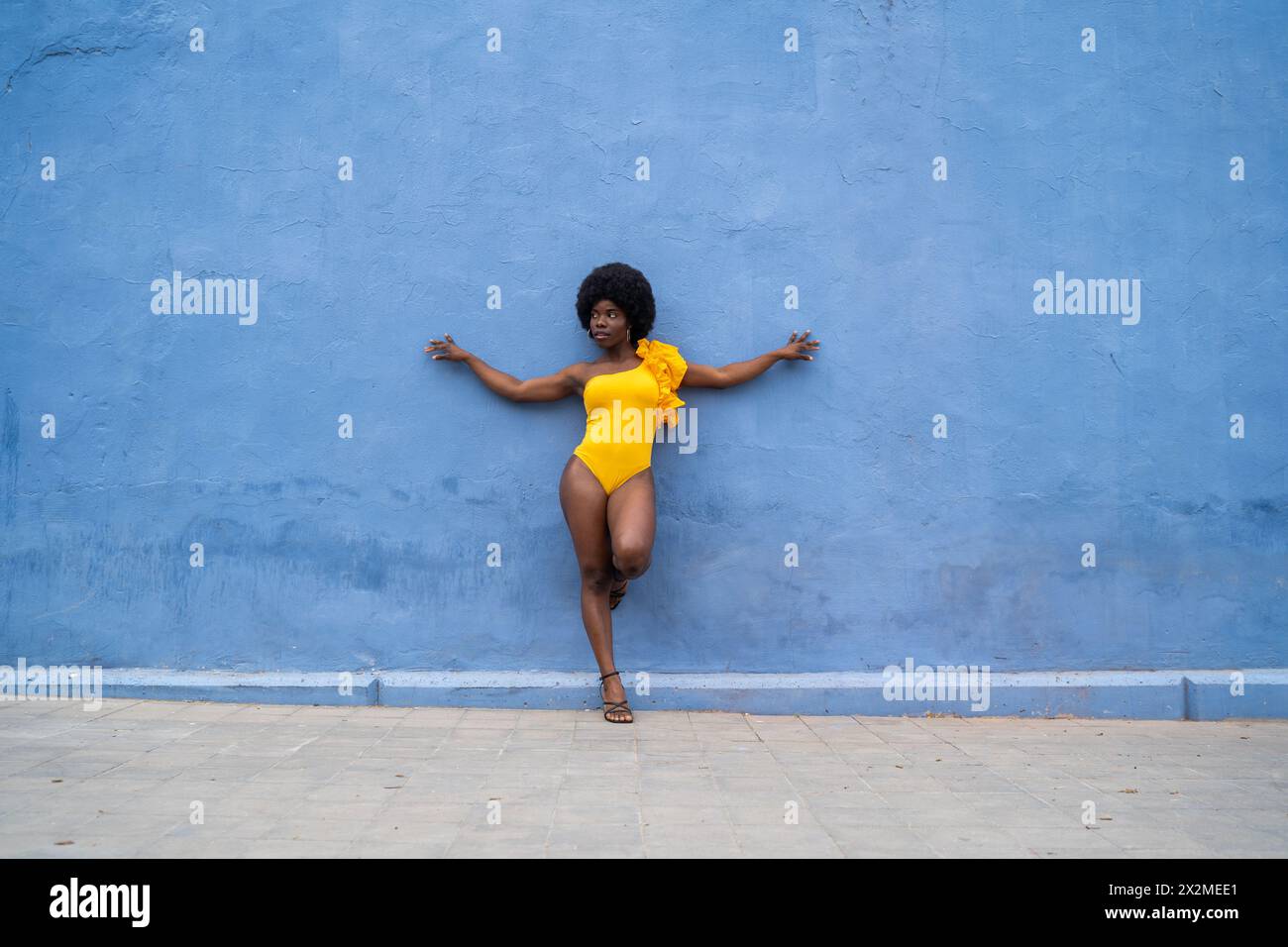 Une femme afro confiante pose dans un monobloc jaune vif sur un fond bleu texturé, dégageant style audacieux et énergie Banque D'Images
