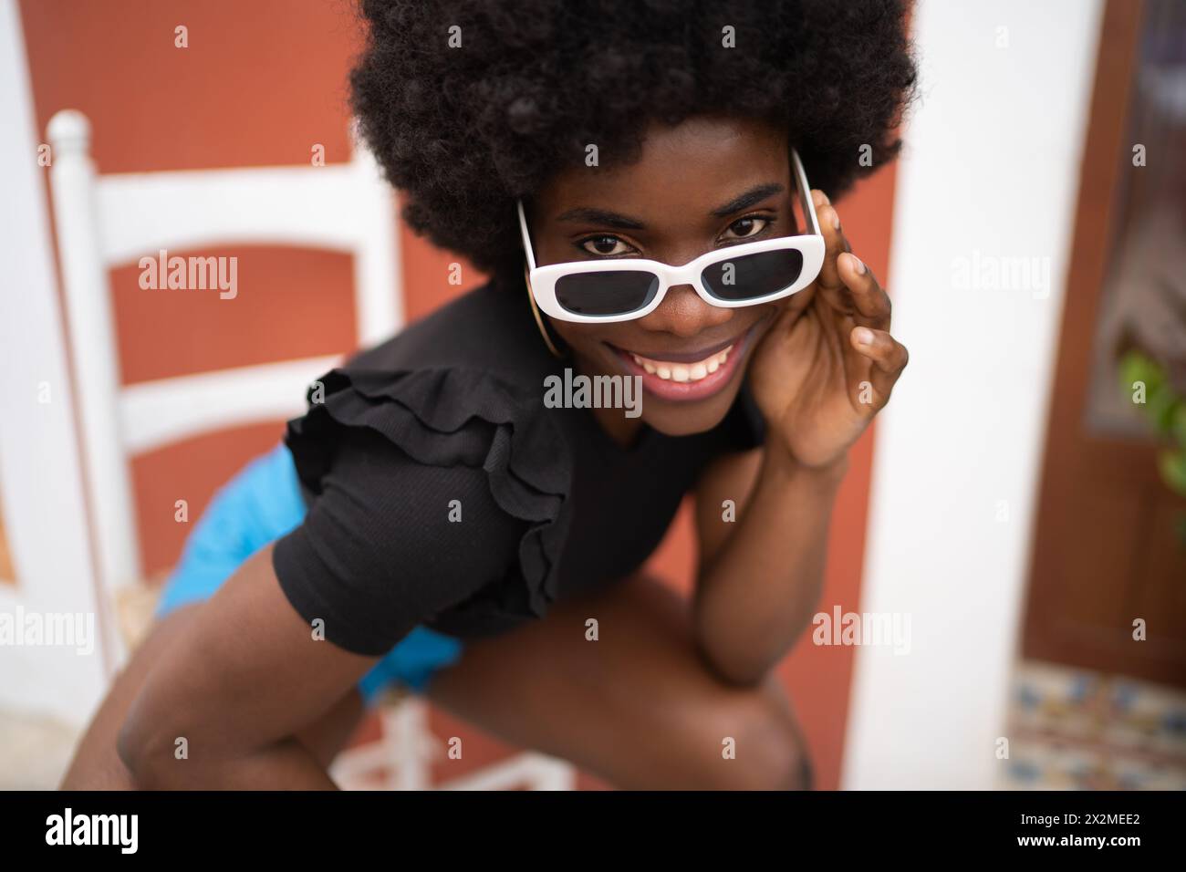 Une jeune femme africaine joyeuse avec une coiffure afro naturelle sourit brillamment tout en ajustant de manière ludique ses lunettes de soleil blanches chics, incarnant un joyeux, Banque D'Images